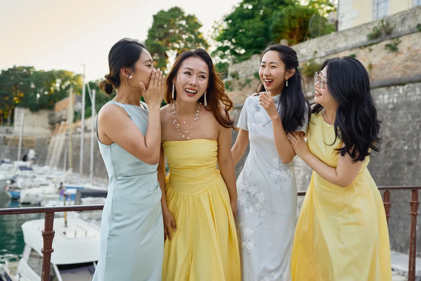 A group of friends in elegant dresses laughing together by the harbour during a photoshoot
